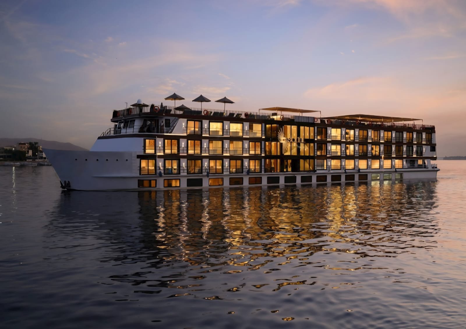 Gran barco de crucero por el Nilo al anochecer, con todas las ventanas de los camarotes iluminadas y su reflejo en el río en calma.