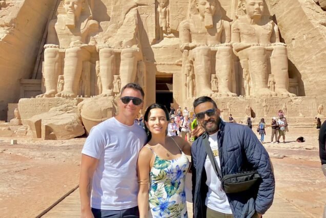 Three travelers standing together in front of the four seated colossi of the Great Temple of Ramesses II at Abu Simbel on a clear day