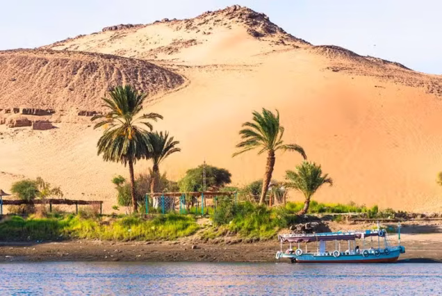 A wide banner crop of the Nile at Aswan, a small covered boat moored below a large orange dune with palms and low green scrub along the shore