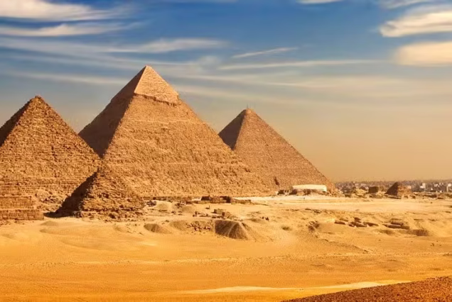 A wide banner crop of the Giza plateau with the three pyramids across open desert and the city faint on the right horizon