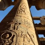Columna monumental de la sala hipostila de Karnak cubierta de jeroglificos y cartuchos faraonicos, vista hacia arriba contra el cielo azul de Egipto