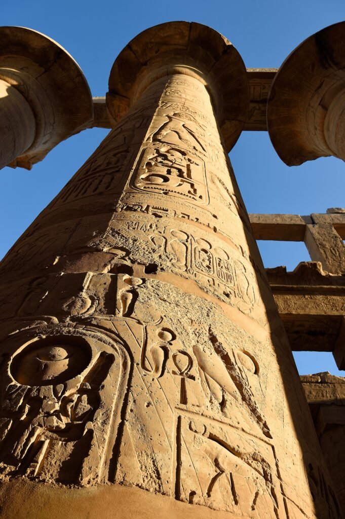 Columna monumental de la sala hipostila de Karnak cubierta de jeroglificos y cartuchos faraonicos, vista hacia arriba contra el cielo azul de Egipto
