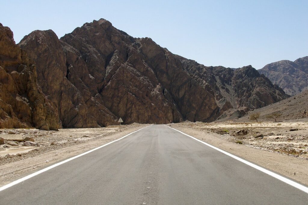 Carretera asfaltada del desierto egipcio que avanza recta hacia montanas escarpadas del Sinai bajo un cielo azul despejado, paisaje ideal para un viaje en coche de alquiler