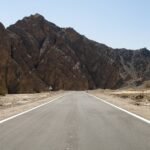 Carretera asfaltada del desierto egipcio que avanza recta hacia montanas escarpadas del Sinai bajo un cielo azul despejado, paisaje ideal para un viaje en coche de alquiler