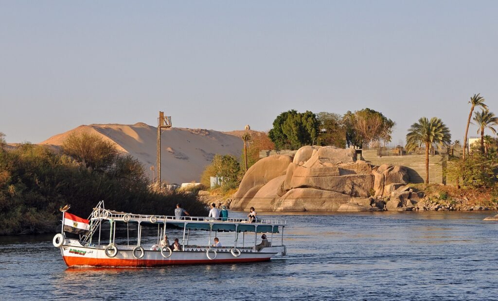 Barco turistico navegando el Nilo al atardecer en Asuan con dunas doradas, rocas de granito y palmeras junto a la isla Elefantina en Egipto