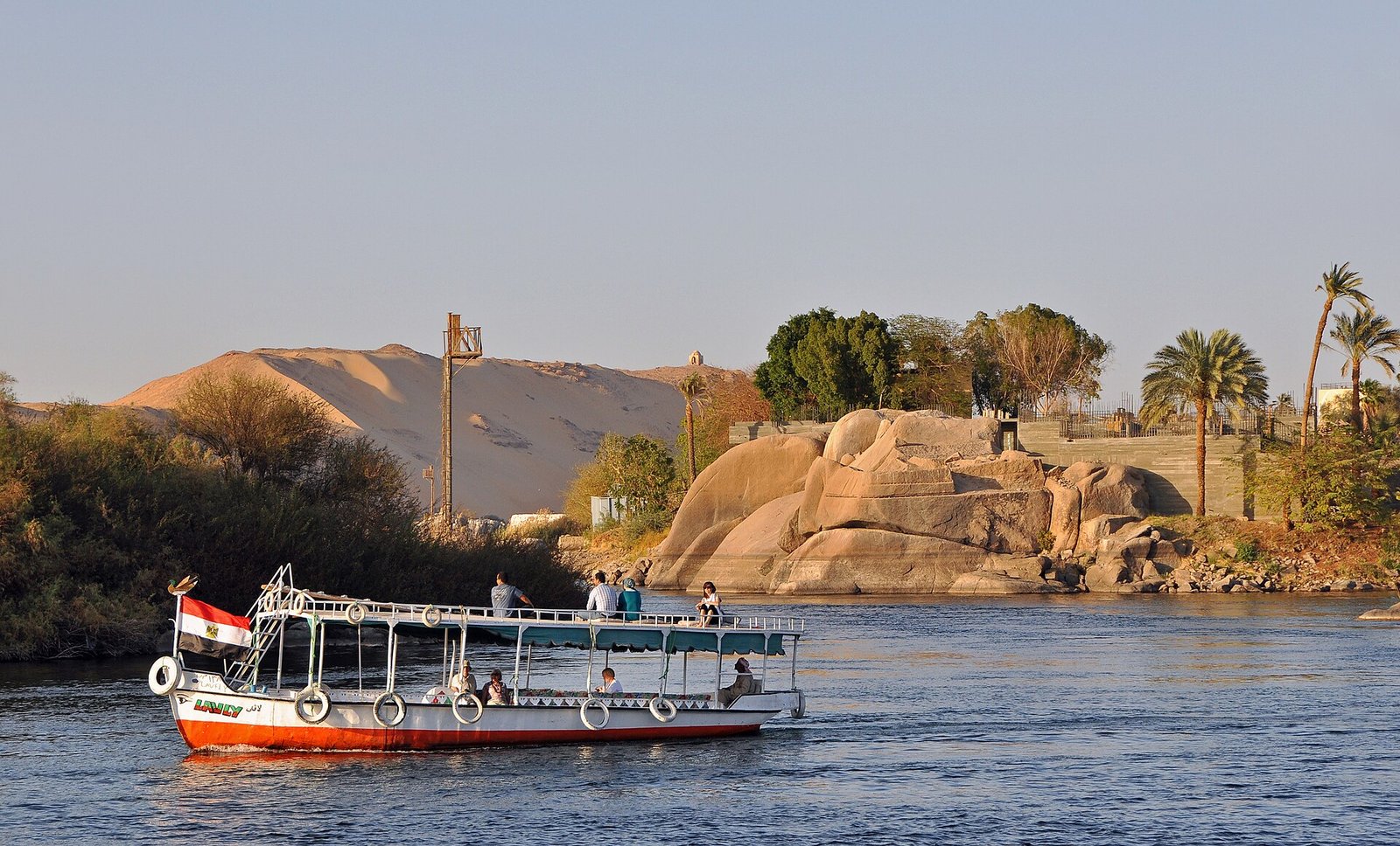 Barco turistico navegando el Nilo al atardecer en Asuan con dunas doradas, rocas de granito y palmeras junto a la isla Elefantina en Egipto