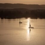 Dos feluccas en silueta navegando el Nilo al atardecer con el reflejo dorado del sol sobre el agua y colinas del desierto al fondo en Egipto