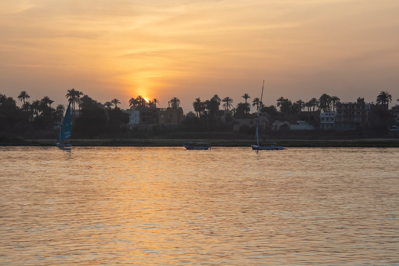 Puesta de sol sobre el Nilo en Egipto con el disco solar dorado tras las palmeras de un pueblo y feluccas tradicionales navegando por el rio