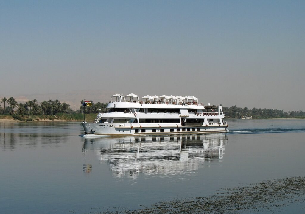 Barco de crucero por el Nilo avanzando sobre aguas tranquilas con su reflejo y palmeras al fondo en Egipto