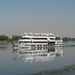 Barco de crucero por el Nilo avanzando sobre aguas tranquilas con su reflejo y palmeras al fondo en Egipto