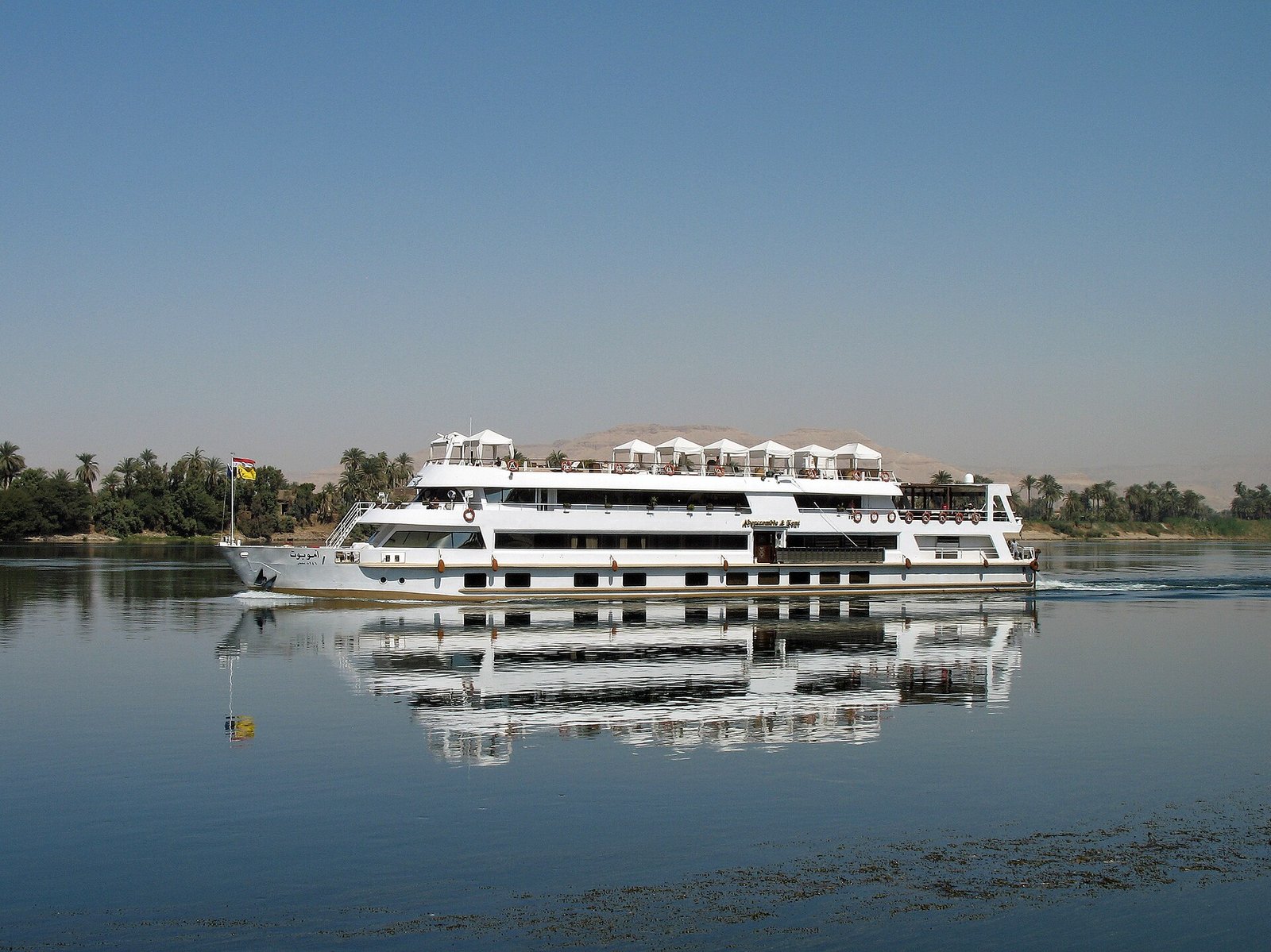 Barco de crucero blanco navegando por el rio Nilo en Egipto con cielo azul y palmeras en la orilla