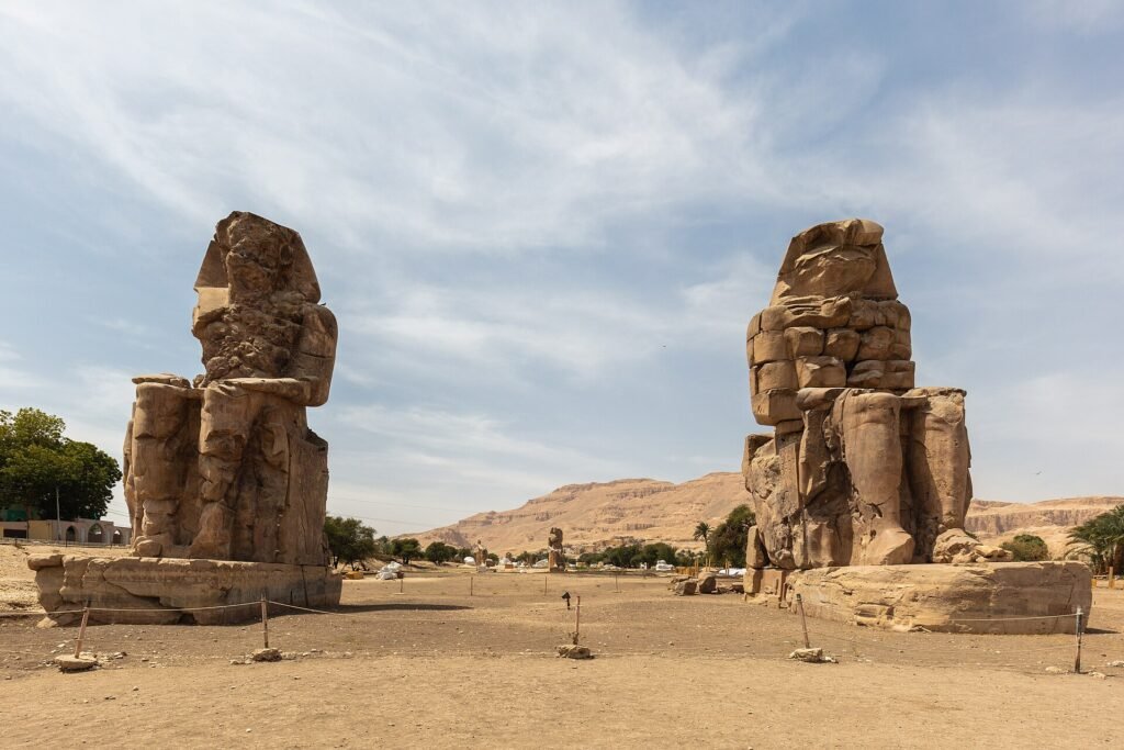 Los dos Colosos de Memnon de piedra erosionada se alzan frente a las colinas tebanas bajo un cielo con nubes en Luxor, Egipto