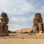 The two Colossi of Memnon standing on the Luxor west bank plain, weathered seated statues with the Theban hills low behind them