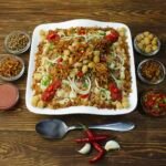 Plato cuadrado blanco de koshari egipcio sobre mesa de madera, con cuencos de cebolla crujiente, garbanzos, lentejas y salsa de tomate alrededor