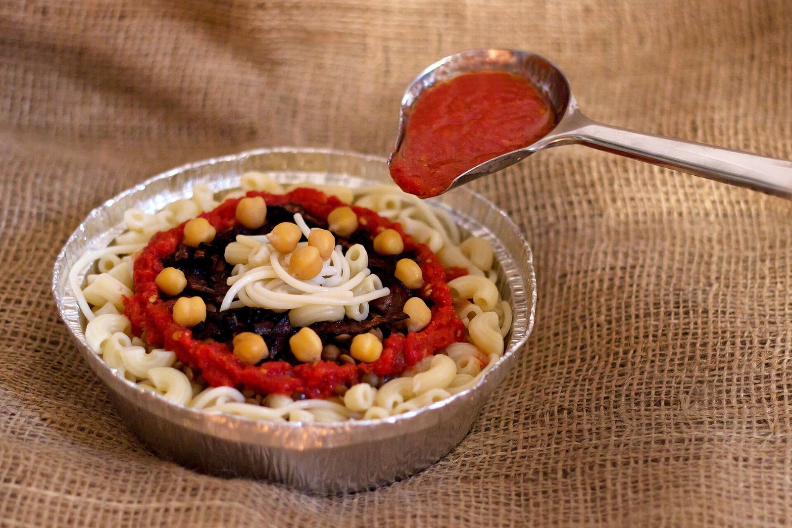 Racion de koshari en recipiente de aluminio sobre arpillera, con macarrones, garbanzos y lentejas, mientras una cuchara vierte salsa de tomate roja encima