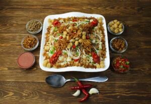 Plato cuadrado blanco de koshari egipcio sobre mesa de madera, con cuencos de cebolla crujiente, garbanzos, lentejas y salsa de tomate alrededor