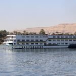 Crucero fluvial blanco navegando por el Nilo bajo cielo azul, con acantilados dorados del desierto y palmeras en la orilla en Egipto