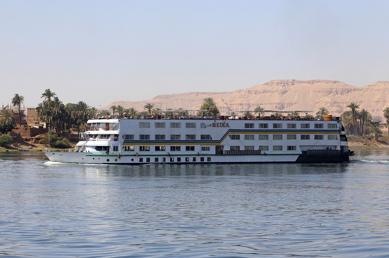 Crucero fluvial blanco navegando por el Nilo bajo cielo azul, con acantilados dorados del desierto y palmeras en la orilla en Egipto