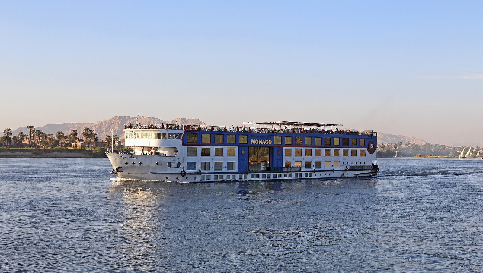 Crucero por el Nilo navegando en aguas tranquilas con luz dorada, montanas y palmeras al fondo y feluccas a vela cerca de Luxor en Egipto