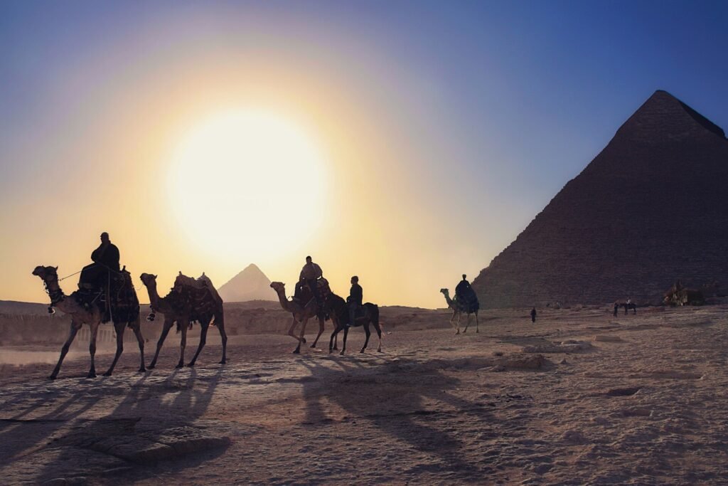 Caravana de camellos con jinetes cruzando el desierto frente a la piramide de Giza al atardecer con un gran sol dorado en Egipto