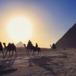 Caravana de camellos con jinetes cruzando el desierto frente a la piramide de Giza al atardecer con un gran sol dorado en Egipto