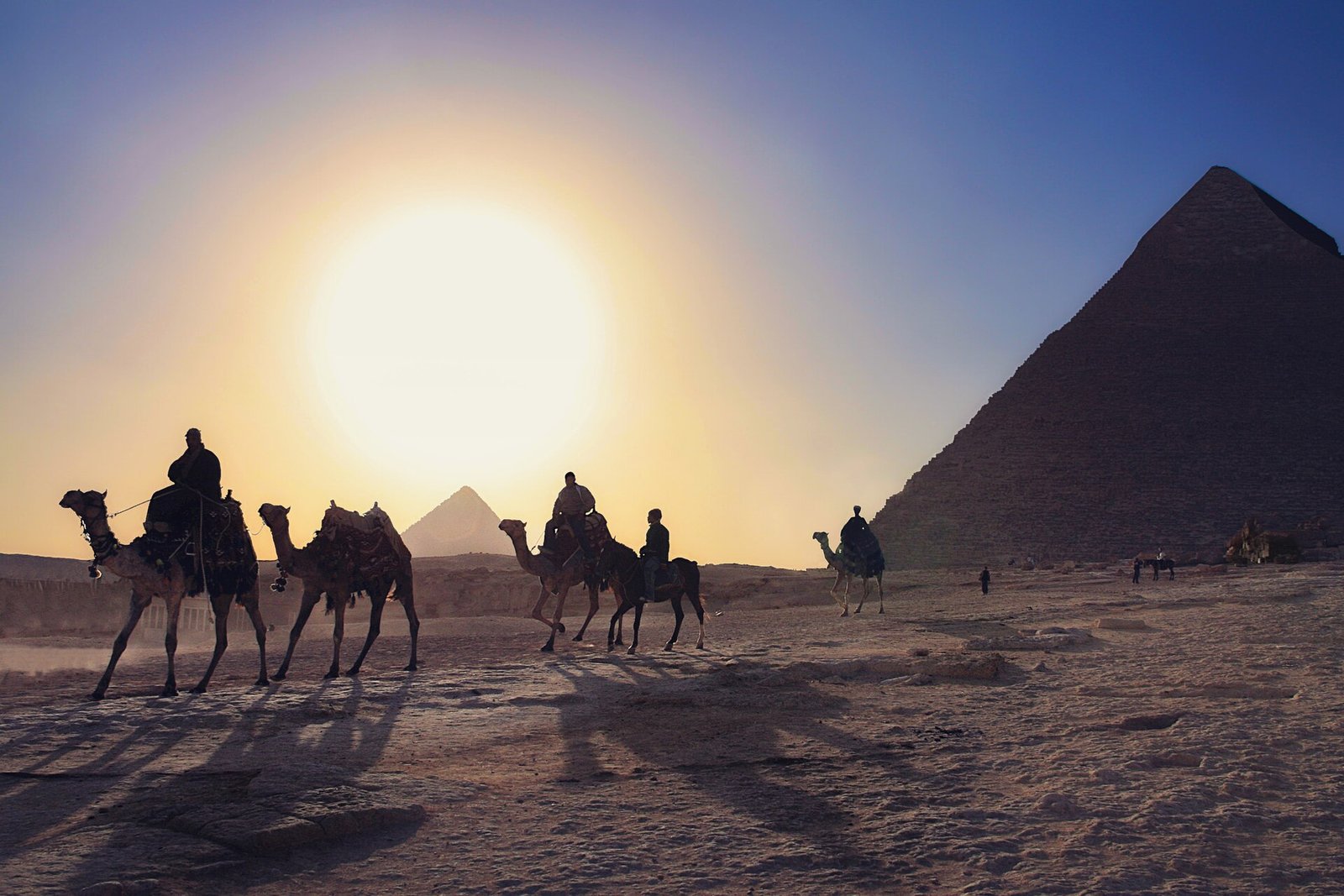 Caravana de camellos con jinetes cruzando el desierto frente a la piramide de Giza al atardecer con un gran sol dorado en Egipto