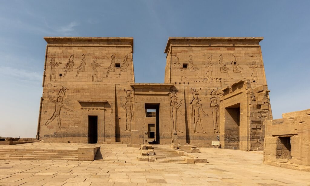 Templo de Philae en Egipto con su gran pilono frontal cubierto de relieves faraonicos, iluminado por luz dorada calida bajo cielo azul, escenario aspiracional