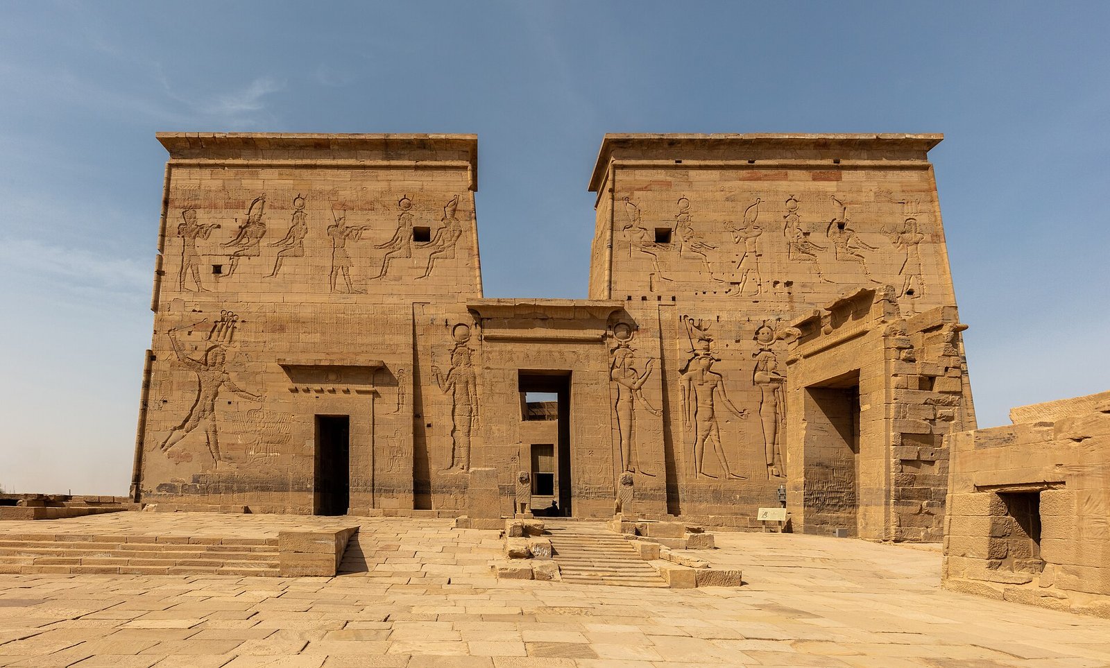 Templo de Philae en Egipto con su gran pilono frontal cubierto de relieves faraonicos, iluminado por luz dorada calida bajo cielo azul, escenario aspiracional