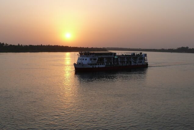 A Nile cruise ship under way at sunset on a wide reach of the river, the sun just above the treeline on the far bank