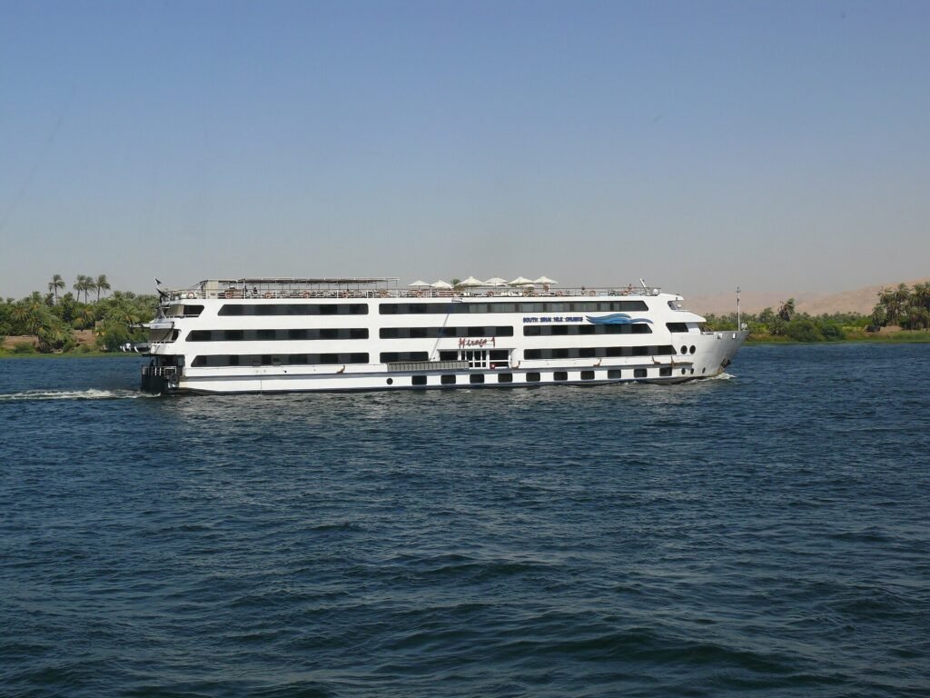 Crucero de lujo por el Nilo, un gran barco blanco de varias cubiertas navegando junto a palmeras y colinas del desierto en Egipto