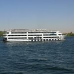 Crucero de lujo por el Nilo, un gran barco blanco de varias cubiertas navegando junto a palmeras y colinas del desierto en Egipto