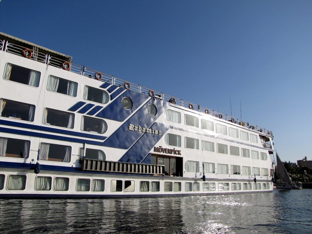 A large white and blue Nile cruise ship photographed close up along its flank, the vessel name Radamis II and a hotel brand mark visible on the hull