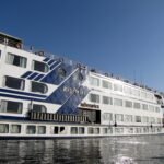 A large white and blue Nile cruise ship photographed close up along its flank, the vessel name Radamis II and a hotel brand mark visible on the hull