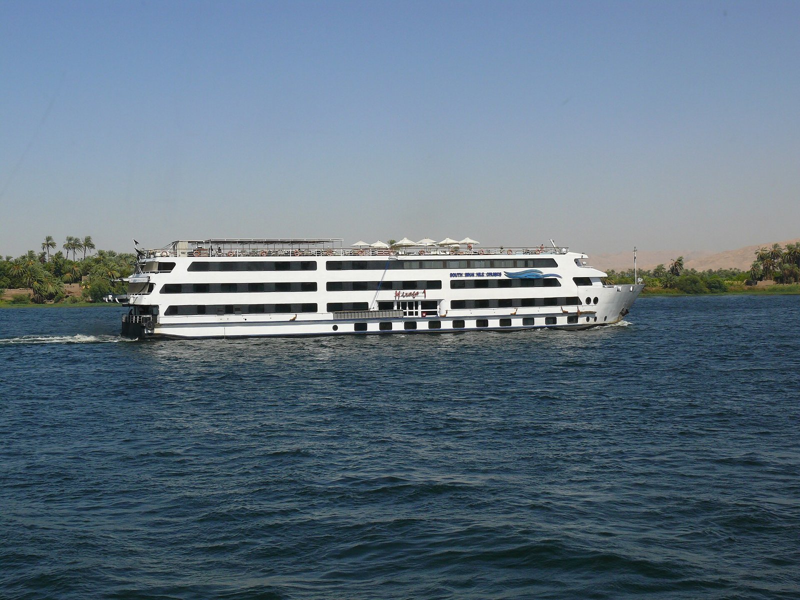 Crucero de lujo por el Nilo, un gran barco blanco de varias cubiertas navegando junto a palmeras y colinas del desierto en Egipto