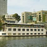 A white two-deck Nile river boat moored alongside a city embankment in daylight, with a canopied top deck, orange life rings along the rail and a gangway to the bank, backed by palms, apartment towers and a green-glass office building; the setting is urban Cairo rather than Upper Egypt.