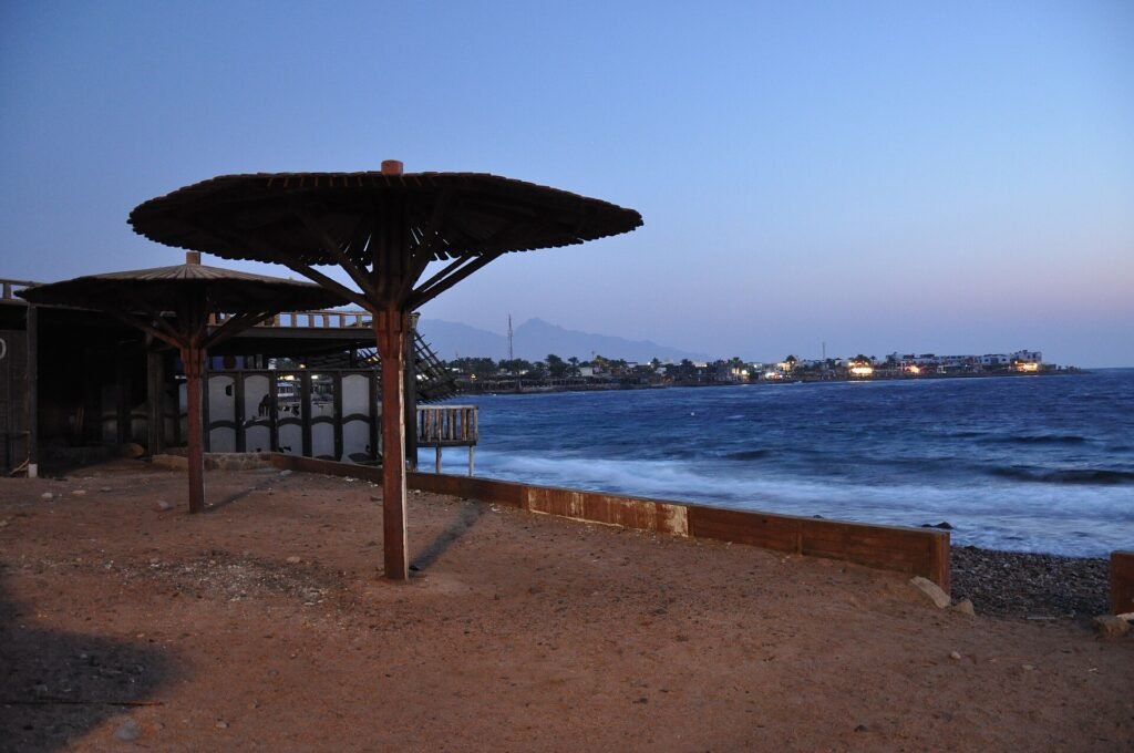 Atardecer en la costa de Dahab en el Sinai con sombrilla beduina de madera, bahia turquesa del Mar Rojo, luces del pueblo y montanas del desierto al fondo en Egipto
