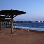 Atardecer en la costa de Dahab en el Sinai con sombrilla beduina de madera, bahia turquesa del Mar Rojo, luces del pueblo y montanas del desierto al fondo en Egipto