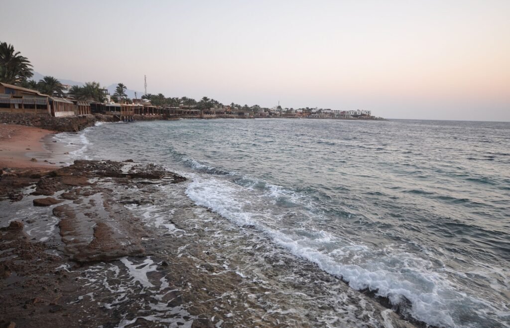Panoramica de la bahia de Dahab al anochecer con olas rompiendo en la orilla rocosa, palmeras y el pueblo costero junto al Mar Rojo en el Sinai, Egipto