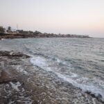 Panoramica de la bahia de Dahab al anochecer con olas rompiendo en la orilla rocosa, palmeras y el pueblo costero junto al Mar Rojo en el Sinai, Egipto