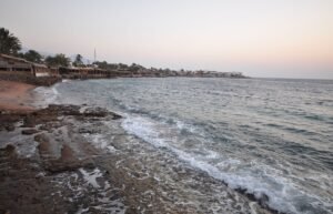 Panoramica de la bahia de Dahab al anochecer con olas rompiendo en la orilla rocosa, palmeras y el pueblo costero junto al Mar Rojo en el Sinai, Egipto