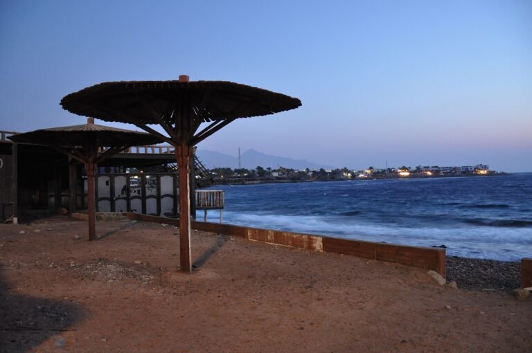 Atardecer en la costa de Dahab en el Sinai con sombrilla beduina de madera, bahia turquesa del Mar Rojo, luces del pueblo y montanas del desierto al fondo en Egipto