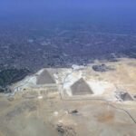 Vista aerea de las tres piramides de Giza sobre la arena dorada con la extensa ciudad de El Cairo al fondo bajo un cielo azul despejado