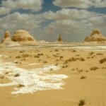 Formaciones de tiza blanca tipo hongo sobre arena dorada ondulada bajo cielo azul con nubes en el Desierto Blanco de Egipto cerca de Farafra