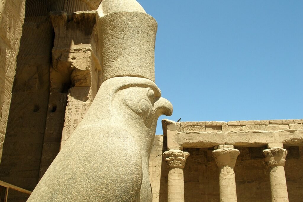 Estatua monumental de granito del dios halcon Horus en el templo de Edfu, con columnas talladas y relieves jeroglificos bajo un cielo azul intenso