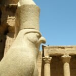 Estatua monumental de granito del dios halcon Horus en el templo de Edfu, con columnas talladas y relieves jeroglificos bajo un cielo azul intenso