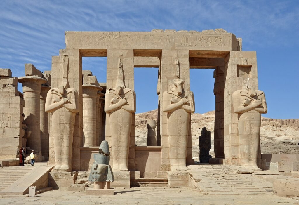 El Ramesseum en Luxor con la columnata de estatuas osiriacas de Ramses II y la cabeza colosal caida bajo un cielo azul en Egipto