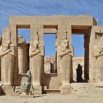 El Ramesseum en Luxor con la columnata de estatuas osiriacas de Ramses II y la cabeza colosal caida bajo un cielo azul en Egipto