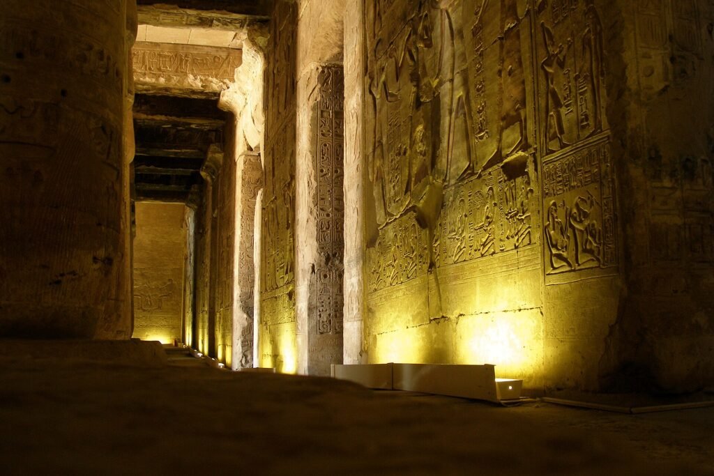 A dim colonnaded hall lit from floor level, deep carved reliefs and hieroglyphs down the wall on the right and a row of square pillars receding into darkness. The temple of Seti I at Abydos