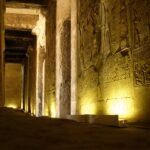 A dim colonnaded hall lit from floor level, deep carved reliefs and hieroglyphs down the wall on the right and a row of square pillars receding into darkness. The temple of Seti I at Abydos