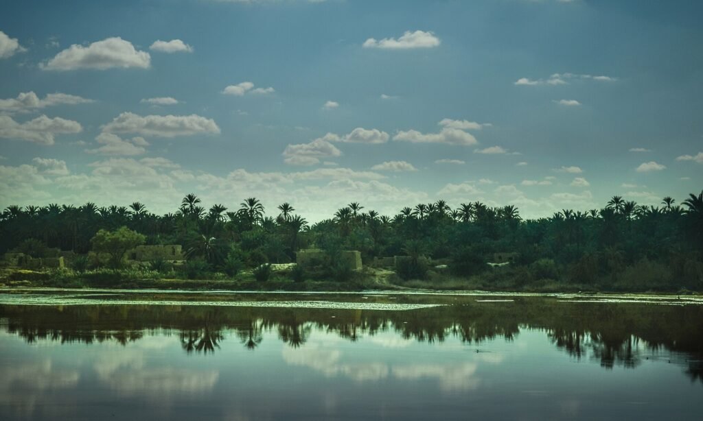 Lago salado turquesa en el oasis de Siwa, Egipto, con el reflejo de palmerales y casas de adobe bajo un cielo con nubes